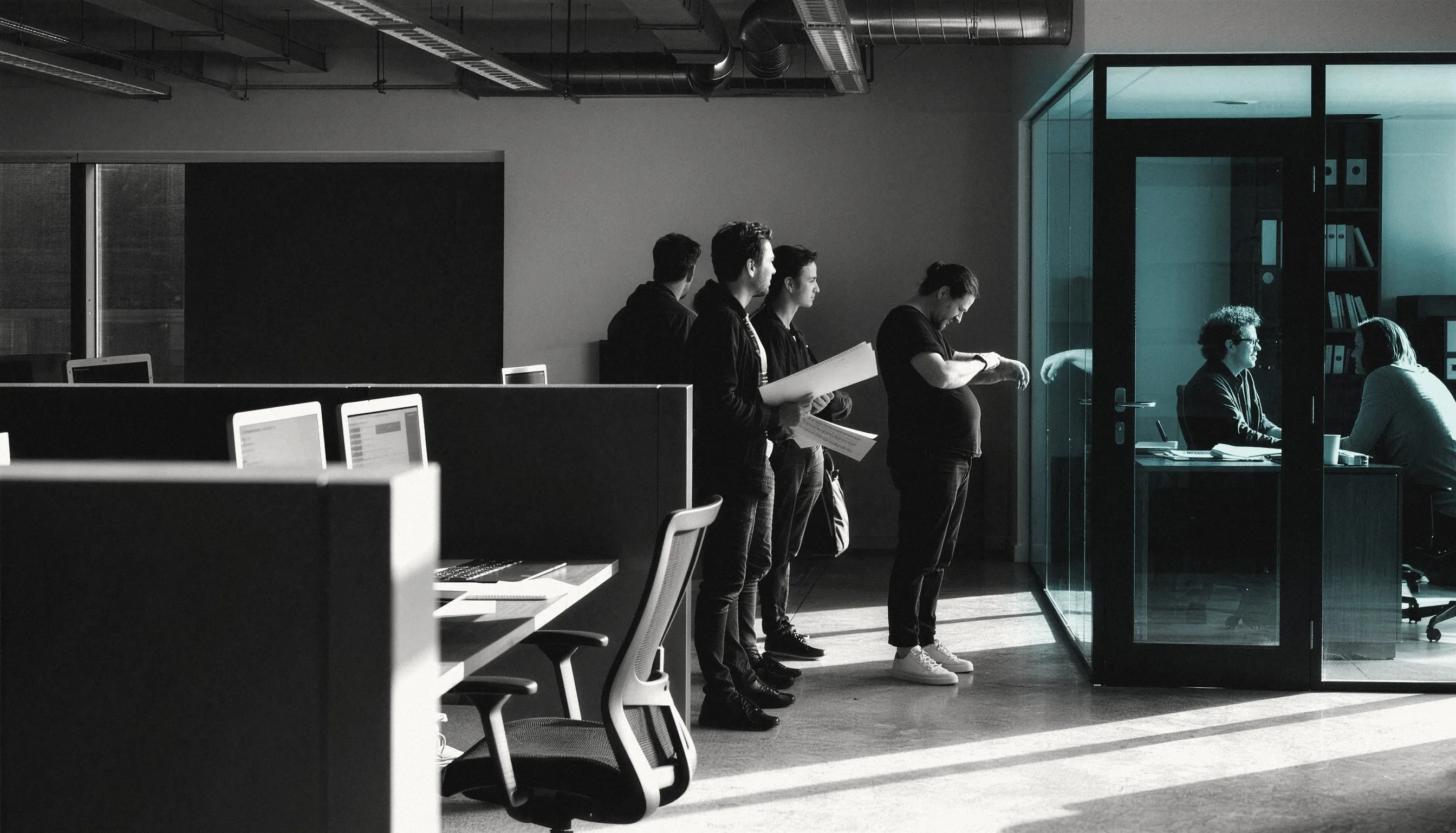 A small glass-walled office: the founder inside with one colleague, and a line of staff waiting outside the door with laptops and paperwork.